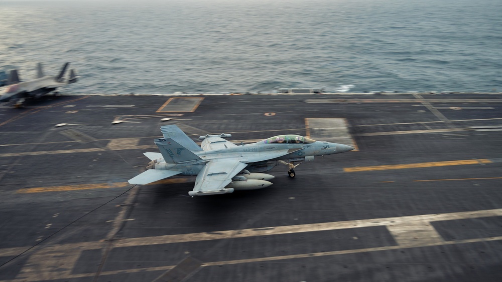 A U.S. Navy jet aircraft, appearing to be an EA-18G Growler or F/A-18F Super Hornet, is captured landing or launching on the flight deck of an aircraft carrier at sea. The aircraft bears the modex number '501' and squadron markings visible on the fuselage. The ocean is visible in the background, indicating open-water carrier operations.