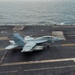 A U.S. Navy jet aircraft, appearing to be an EA-18G Growler or F/A-18F Super Hornet, is captured landing or launching on the flight deck of an aircraft carrier at sea. The aircraft bears the modex number '501' and squadron markings visible on the fuselage. The ocean is visible in the background, indicating open-water carrier operations.