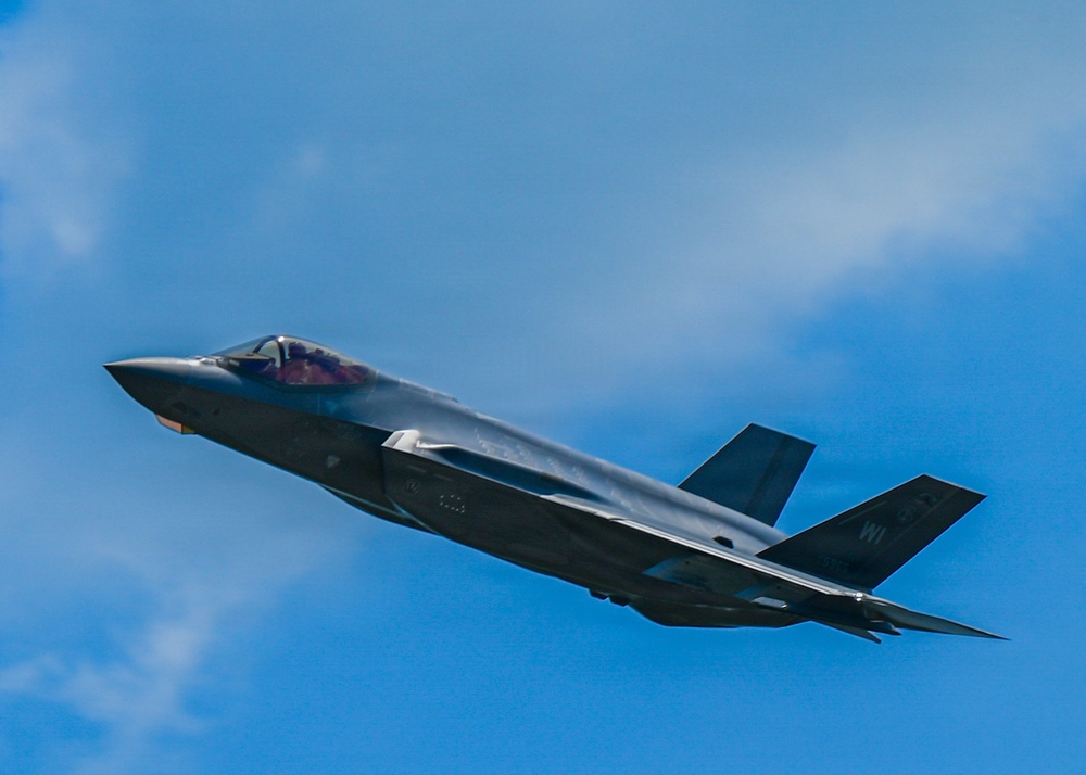 A single-seat stealth fighter jet, identifiable as an F-35 Lightning II, is captured in flight against a partly cloudy blue sky. The aircraft is shown in a slight banking maneuver, revealing its distinctive delta-wing design and twin tail fins. Tail markings include the letters 'WI', suggesting a Wisconsin Air National Guard unit.