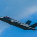 A single-seat stealth fighter jet, identifiable as an F-35 Lightning II, is captured in flight against a partly cloudy blue sky. The aircraft is shown in a slight banking maneuver, revealing its distinctive delta-wing design and twin tail fins. Tail markings include the letters 'WI', suggesting a Wisconsin Air National Guard unit.