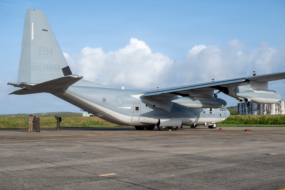 A U.S. Marine Corps C-130J Super Hercules transport aircraft is parked on a tarmac, identifiable by the tail code 'BH' and serial number '9069'. The aircraft is painted in standard gray military livery and appears to be on a flight line at a tropical or subtropical airfield. Several personnel in military uniforms are standing near the left side of the aircraft.