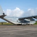 A U.S. Marine Corps C-130J Super Hercules transport aircraft is parked on a tarmac, identifiable by the tail code 'BH' and serial number '9069'. The aircraft is painted in standard gray military livery and appears to be on a flight line at a tropical or subtropical airfield. Several personnel in military uniforms are standing near the left side of the aircraft.