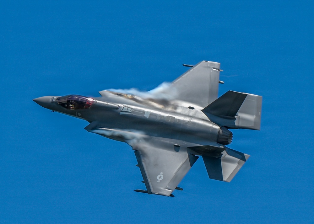 A single-engine stealth fighter jet, consistent with an F-35 Lightning II, is captured in low-altitude high-speed flight against a clear blue sky. The aircraft is banked at an angle, revealing its top surface, with visible vapor condensation forming over the wings and fuselage due to aerodynamic pressure changes. A small roundel insignia is visible on the lower fuselage, suggesting a military operator.