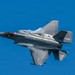 A single-engine stealth fighter jet, consistent with an F-35 Lightning II, is captured in low-altitude high-speed flight against a clear blue sky. The aircraft is banked at an angle, revealing its top surface, with visible vapor condensation forming over the wings and fuselage due to aerodynamic pressure changes. A small roundel insignia is visible on the lower fuselage, suggesting a military operator.