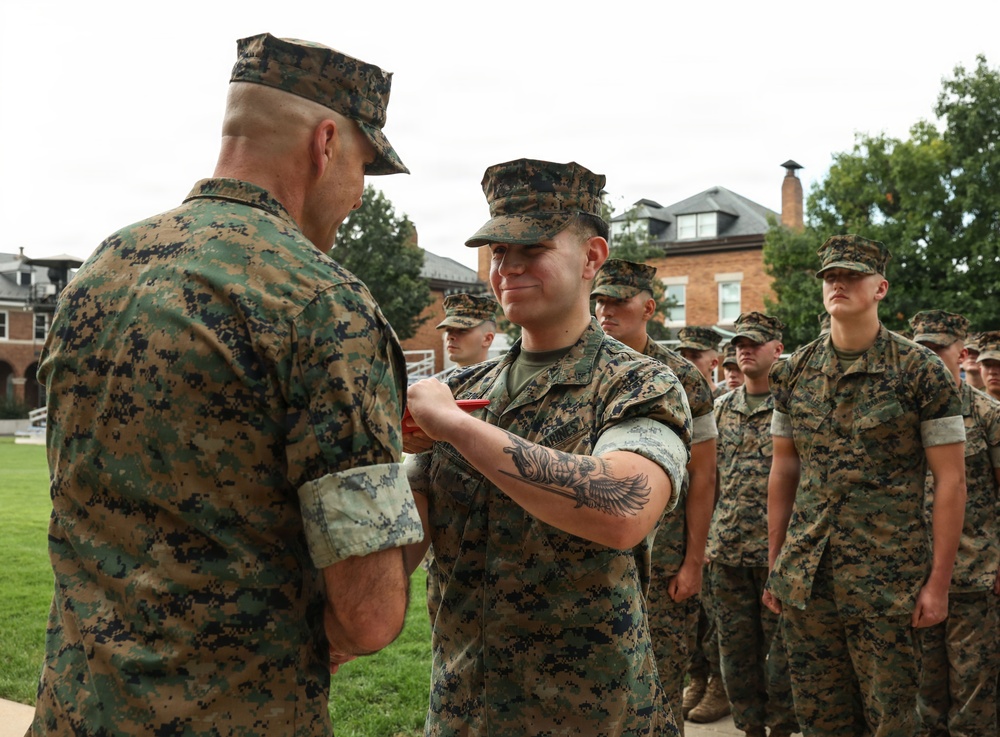 A military ceremony is taking place outdoors on what appears to be a base or installation. A senior Marine is pinning a medal or award onto a junior Marine who is smiling, while a formation of Marines stands at attention in the background. All personnel are wearing MARPAT (Marine Pattern) camouflage uniforms.