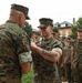 A military ceremony is taking place outdoors on what appears to be a base or installation. A senior Marine is pinning a medal or award onto a junior Marine who is smiling, while a formation of Marines stands at attention in the background. All personnel are wearing MARPAT (Marine Pattern) camouflage uniforms.