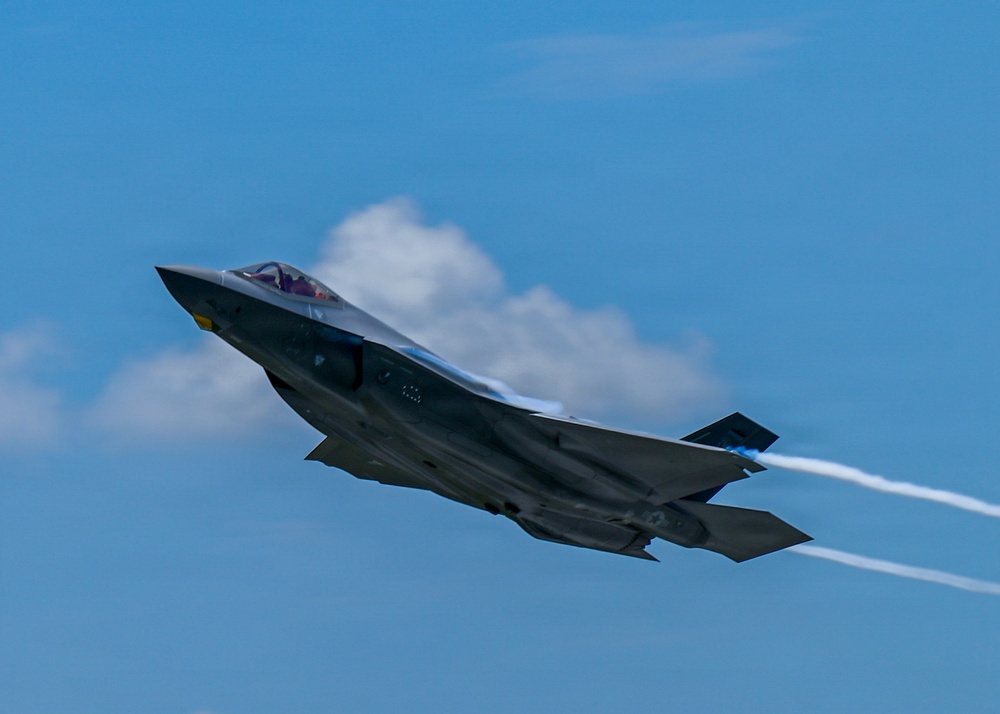 A single-seat stealth fighter jet, consistent with an F-35 Lightning II, is captured in flight against a blue sky with scattered clouds. The aircraft is in a climbing or banking maneuver, producing a visible exhaust contrail from its single engine. The image is sharp and well-composed, suggesting a professional airshow or demonstration photography setting.