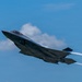 A single-seat stealth fighter jet, consistent with an F-35 Lightning II, is captured in flight against a blue sky with scattered clouds. The aircraft is in a climbing or banking maneuver, producing a visible exhaust contrail from its single engine. The image is sharp and well-composed, suggesting a professional airshow or demonstration photography setting.
