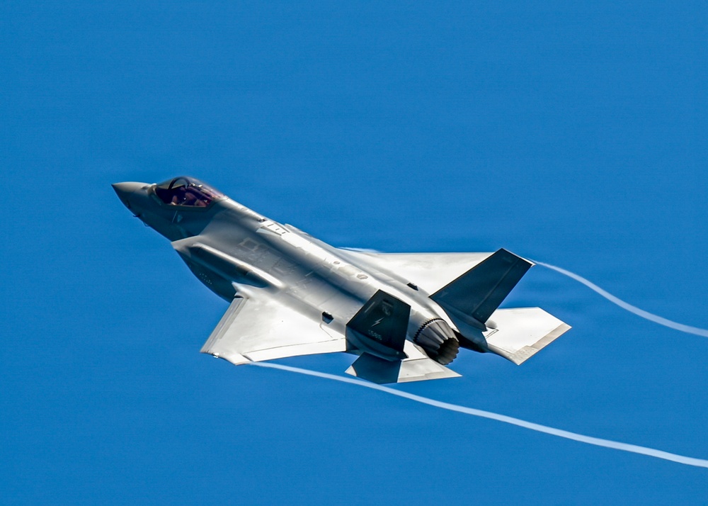 A single-engine stealth multirole fighter jet, consistent with an F-35 Lightning II, is captured in flight against a clear blue sky. The aircraft is banking slightly and generating wingtip/wing vortex condensation trails. The image is a high-quality, close-range aerial photograph showing significant detail of the aircraft's airframe.