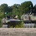 The image shows two wheeled armored military vehicles in woodland camouflage pattern positioned side by side in an open field or training area. Both vehicles are equipped with green signal flags, antennas, and various mounted equipment on their turrets. The setting appears to be a military exercise or training operation, with dense pine forest visible in the background.