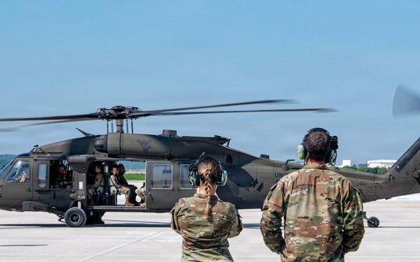 A military UH-60 Black Hawk helicopter is on a tarmac with its rotors spinning, observed by two soldiers standing with their backs to the camera. A third soldier is walking away from the aircraft on the left side. The scene appears to be a routine ground operation or landing procedure at a military airfield.