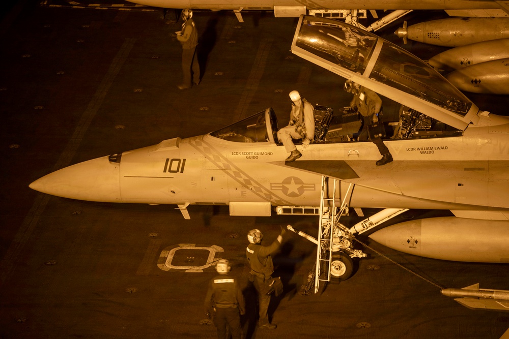An aerial night-time view of a U.S. Navy fighter jet (number 101) on an aircraft carrier deck, illuminated by warm artificial lighting. The aircraft is a two-seat variant, likely an F/A-18F Super Hornet, with pilot names 'LCDR SCOTT GUIMOND / GUIDO' and 'LCDR WILLIAM EWALD / WALDO' stenciled on the fuselage. Ground crew and flight personnel are actively engaged in pre-flight or post-flight operations around the aircraft.