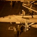 An aerial night-time view of a U.S. Navy fighter jet (number 101) on an aircraft carrier deck, illuminated by warm artificial lighting. The aircraft is a two-seat variant, likely an F/A-18F Super Hornet, with pilot names 'LCDR SCOTT GUIMOND / GUIDO' and 'LCDR WILLIAM EWALD / WALDO' stenciled on the fuselage. Ground crew and flight personnel are actively engaged in pre-flight or post-flight operations around the aircraft.