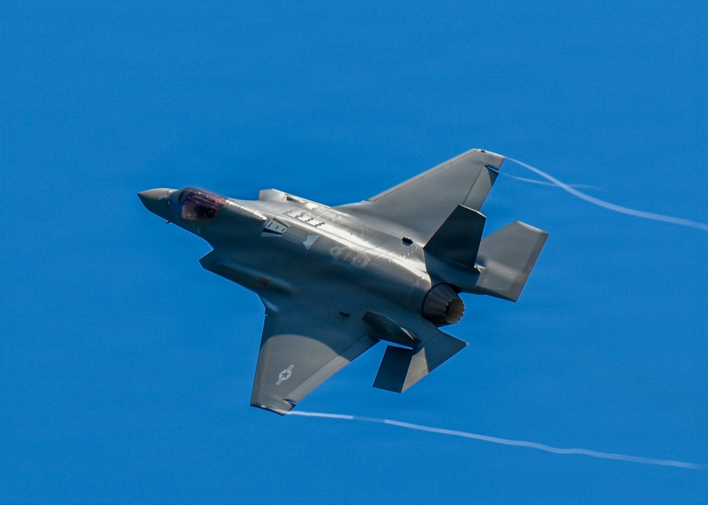 A single-engine stealth fighter jet, identifiable as an F-35 Lightning II, is captured in flight against a clear blue sky. The aircraft is shown in a banked maneuver, revealing its top surface, with visible wingtip vortex condensation trails streaming from the wings. The gray paint scheme and distinctive delta-wing design are clearly visible.