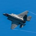 A single-engine stealth fighter jet, identifiable as an F-35 Lightning II, is captured in flight against a clear blue sky. The aircraft is shown in a banked maneuver, revealing its top surface, with visible wingtip vortex condensation trails streaming from the wings. The gray paint scheme and distinctive delta-wing design are clearly visible.