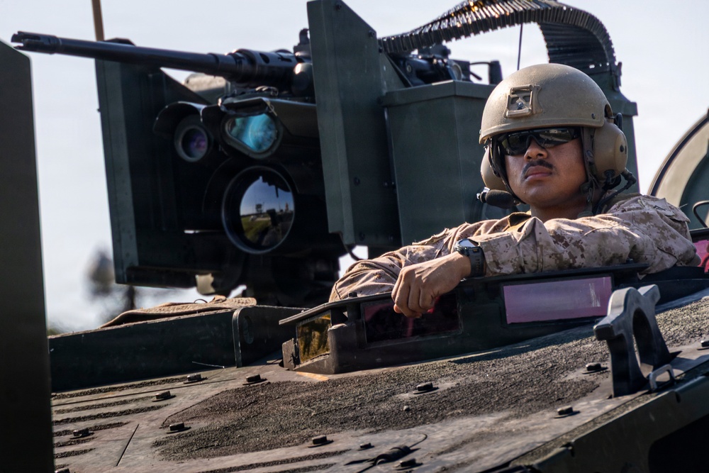 A military service member wearing desert camouflage (MARPAT pattern) and a tan combat helmet is positioned in the hatch of an armored vehicle. The soldier is equipped with communications headgear and sunglasses, resting their arms on the vehicle's turret ring. Behind them, a mounted machine gun with an ammunition belt and optical/targeting systems are clearly visible.