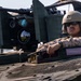 A military service member wearing desert camouflage (MARPAT pattern) and a tan combat helmet is positioned in the hatch of an armored vehicle. The soldier is equipped with communications headgear and sunglasses, resting their arms on the vehicle's turret ring. Behind them, a mounted machine gun with an ammunition belt and optical/targeting systems are clearly visible.