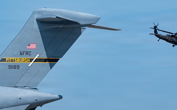 The image shows the tail section of a U.S. Air Force Reserve Command (AFRC) fixed-wing aircraft assigned to Pittsburgh, with tail number 91189, on a tarmac. In the background, a military helicopter — consistent with a UH-60 Black Hawk — is airborne near what appear to be large aircraft hangars. The scene depicts routine military airfield operations at what is likely Pittsburgh International Airport Air Reserve Station.