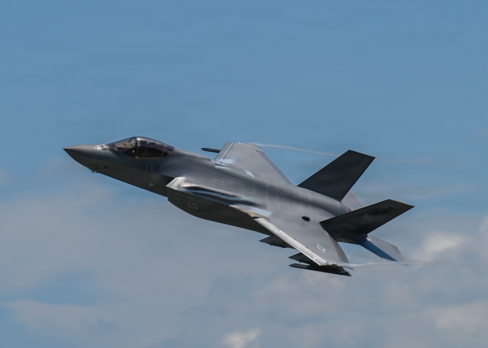 A single-seat stealth fighter jet, consistent with an F-35 Lightning II, is captured in low-altitude flight against a partly cloudy blue sky. The aircraft is in a slight bank, revealing its underside and characteristic delta-wing design. Aerodynamic vapor condensation is visible streaming off the fuselage, indicating high-speed or high-G maneuvering.