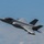 The F-35A Lightning II offically arrives at the 104th Fighter Wing, brings new era to Barnes Air National Guard Base A single-seat stealth fighter jet, consistent with an F-35 Lightning II, is captured in low-altitude flight against a partly cloudy blue sky. The aircraft is in a slight bank, revealing its underside and characteristic delta-wing design. Aerodynamic vapor condensation is visible streaming off the fuselage, indicating high-speed or high-G maneuvering.