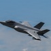 A single-seat stealth fighter jet, consistent with an F-35 Lightning II, is captured in low-altitude flight against a partly cloudy blue sky. The aircraft is in a slight bank, revealing its underside and characteristic delta-wing design. Aerodynamic vapor condensation is visible streaming off the fuselage, indicating high-speed or high-G maneuvering.