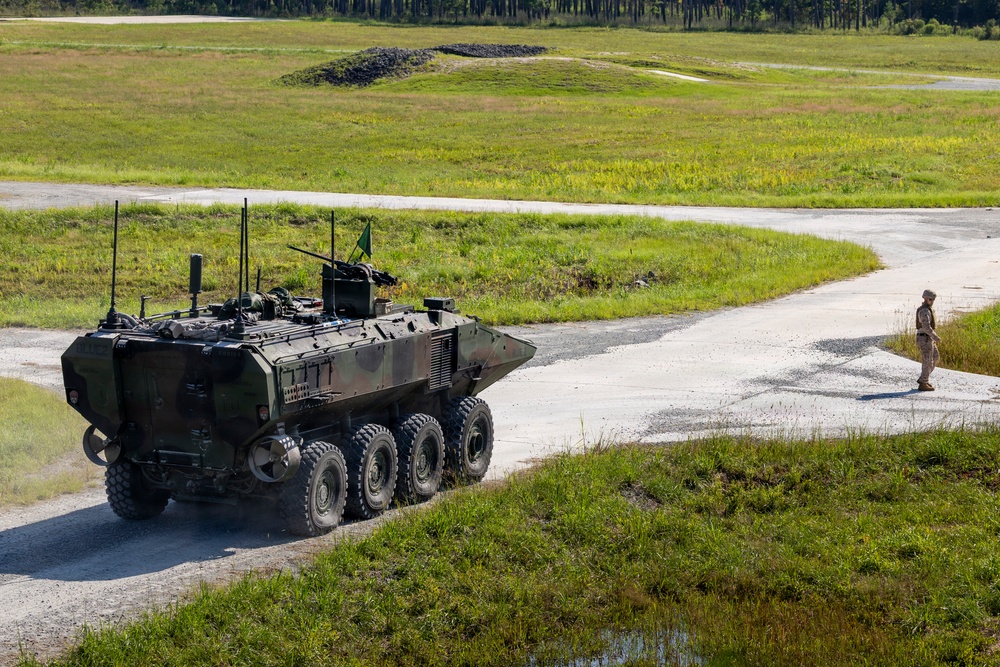 A large 8-wheeled armored military vehicle in camouflage paint is traveling along a paved road on what appears to be a military training range or installation. A single soldier in tan/khaki uniform stands on the road ahead of the vehicle, possibly acting as a guide or observer. The surrounding terrain consists of open grassland with tree lines visible in the background.