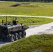 A large 8-wheeled armored military vehicle in camouflage paint is traveling along a paved road on what appears to be a military training range or installation. A single soldier in tan/khaki uniform stands on the road ahead of the vehicle, possibly acting as a guide or observer. The surrounding terrain consists of open grassland with tree lines visible in the background.