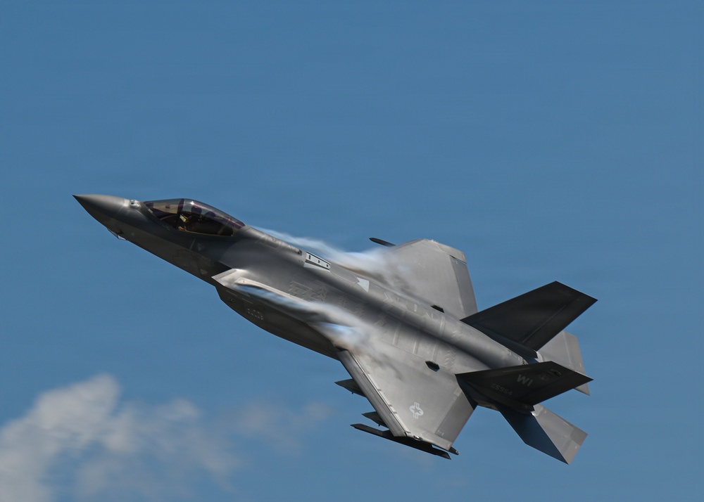 A single-seat stealth fighter jet, consistent with an F-35 Lightning II, is captured in flight against a clear blue sky. The aircraft is banking at an angle, revealing its top surface, with visible vapor condensation forming over the fuselage and wings, indicating high-speed maneuvering. The image appears to have been taken at an airshow or during a demonstration flight.