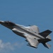 A single-seat stealth fighter jet, consistent with an F-35 Lightning II, is captured in flight against a clear blue sky. The aircraft is banking at an angle, revealing its top surface, with visible vapor condensation forming over the fuselage and wings, indicating high-speed maneuvering. The image appears to have been taken at an airshow or during a demonstration flight.