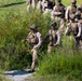 A group of military personnel in full combat gear are moving through a grassy, vegetated area along what appears to be a dirt or gravel path. The soldiers are wearing desert/woodland camouflage uniforms, tactical vests, helmets, and are carrying rifles. This appears to be a training exercise or patrol operation conducted in a field environment.