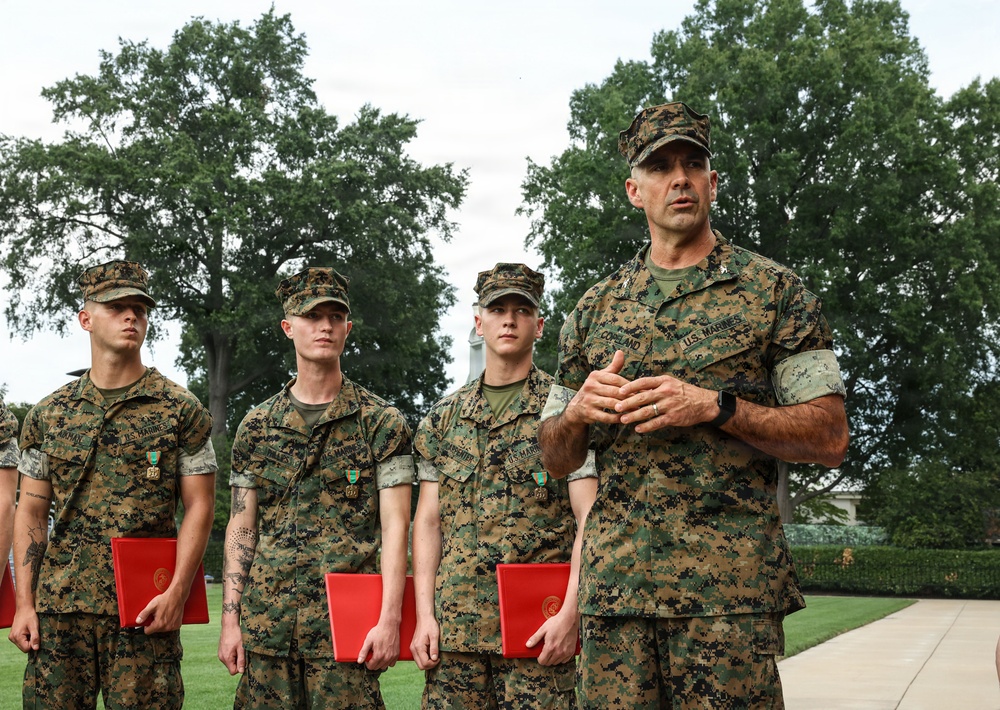 A group of U.S. Marines in MARPAT woodland camouflage uniforms are gathered outdoors on a manicured lawn, apparently during an awards or recognition ceremony. Three junior Marines are holding red folders and wearing medals on their chests, while a senior Marine (name tape reads 'COPELAND') addresses them with hands clasped. The setting appears to be a formal military installation with large trees and a white building visible in the background.