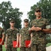 A group of U.S. Marines in MARPAT woodland camouflage uniforms are gathered outdoors on a manicured lawn, apparently during an awards or recognition ceremony. Three junior Marines are holding red folders and wearing medals on their chests, while a senior Marine (name tape reads 'COPELAND') addresses them with hands clasped. The setting appears to be a formal military installation with large trees and a white building visible in the background.