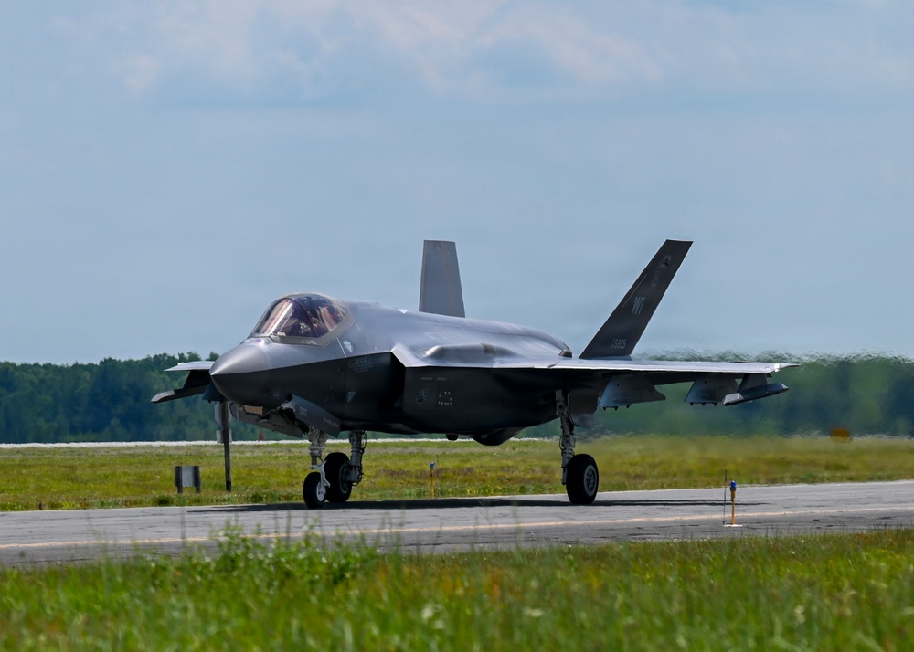A fifth-generation stealth fighter jet, identifiable as an F-35A Lightning II, is taxiing on a runway at a military airbase. The aircraft is viewed from a front-quarter angle, showing its distinctive delta-wing design and twin tail fins. The setting appears to be a well-maintained airfield surrounded by green vegetation under partly cloudy skies.