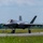 The F-35A Lightning II offically arrives at the 104th Fighter Wing, brings new era to Barnes Air National Guard Base A fifth-generation stealth fighter jet, identifiable as an F-35A Lightning II, is taxiing on a runway at a military airbase. The aircraft is viewed from a front-quarter angle, showing its distinctive delta-wing design and twin tail fins. The setting appears to be a well-maintained airfield surrounded by green vegetation under partly cloudy skies.