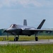 A fifth-generation stealth fighter jet, identifiable as an F-35A Lightning II, is taxiing on a runway at a military airbase. The aircraft is viewed from a front-quarter angle, showing its distinctive delta-wing design and twin tail fins. The setting appears to be a well-maintained airfield surrounded by green vegetation under partly cloudy skies.