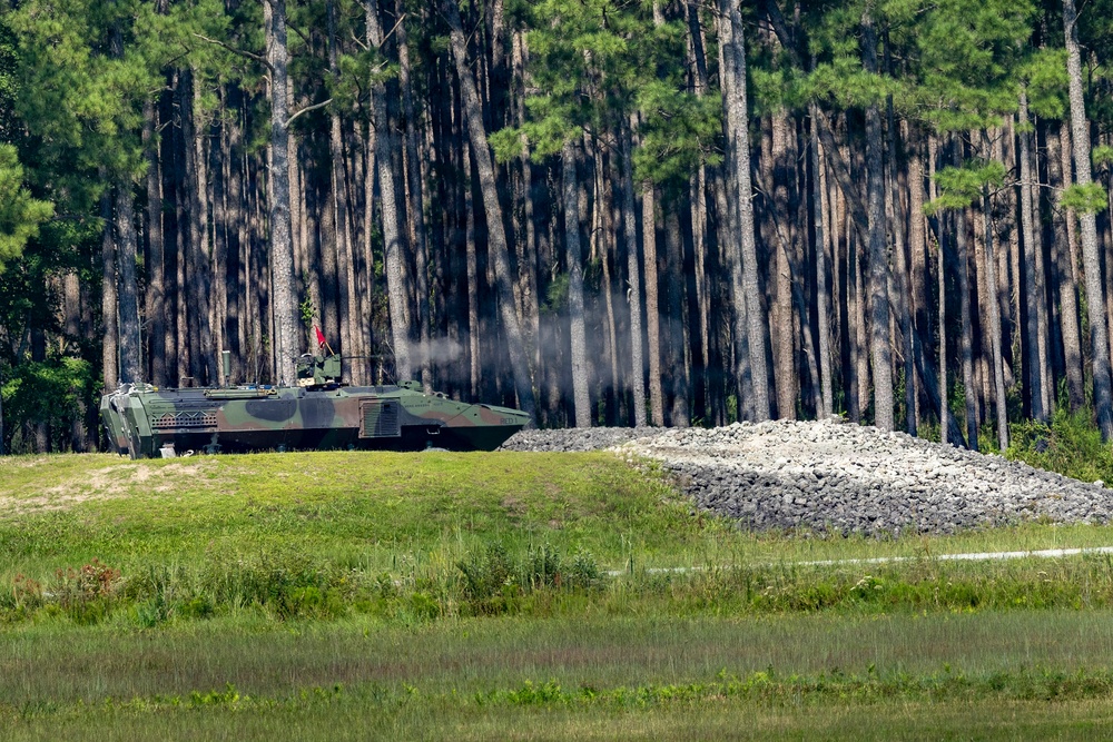 A camouflage-painted armored fighting vehicle is positioned near a treeline of tall pine trees, with visible smoke or exhaust rising from the vehicle. A large pile of gravel or crushed rock is situated to the right of the vehicle. The setting appears to be a military training range or exercise area.