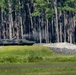 A camouflage-painted armored fighting vehicle is positioned near a treeline of tall pine trees, with visible smoke or exhaust rising from the vehicle. A large pile of gravel or crushed rock is situated to the right of the vehicle. The setting appears to be a military training range or exercise area.