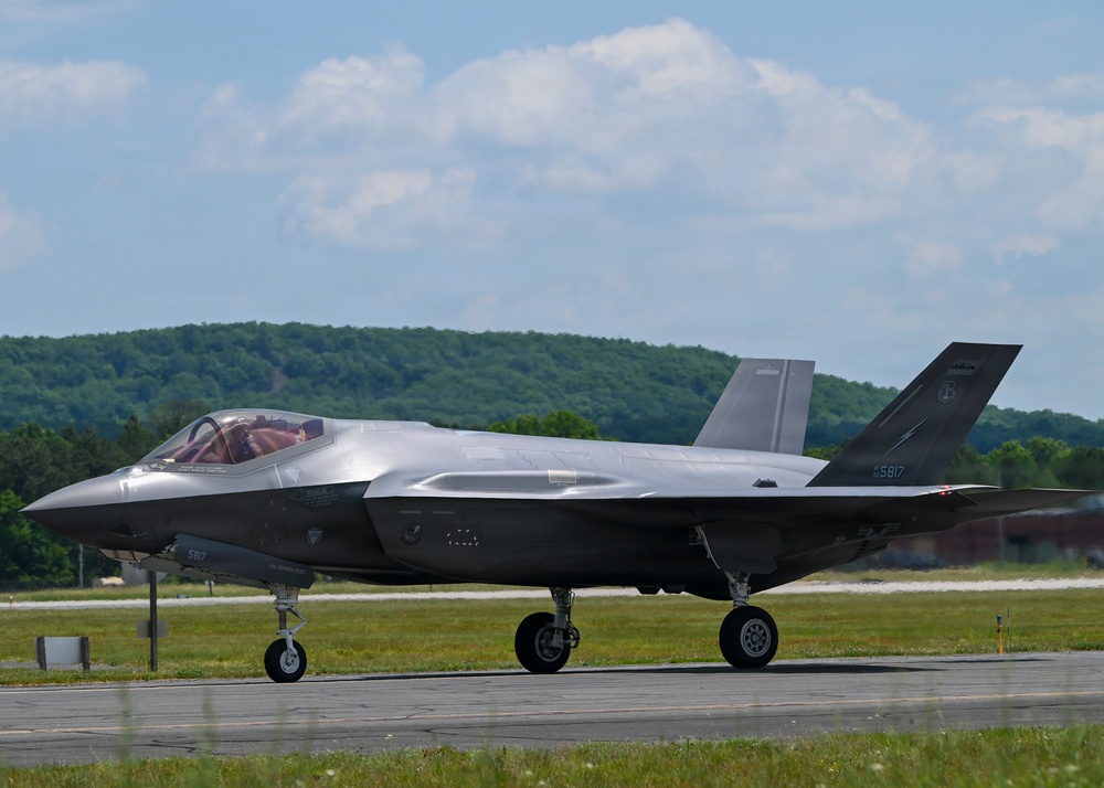 A single F-35 Lightning II stealth fighter jet is taxiing on a runway at a military airfield. The aircraft bears the tail number '5817' and has a pilot visible in the cockpit. The setting appears to be a warm, clear day with green wooded hills visible in the background.
