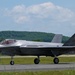 A single F-35 Lightning II stealth fighter jet is taxiing on a runway at a military airfield. The aircraft bears the tail number '5817' and has a pilot visible in the cockpit. The setting appears to be a warm, clear day with green wooded hills visible in the background.