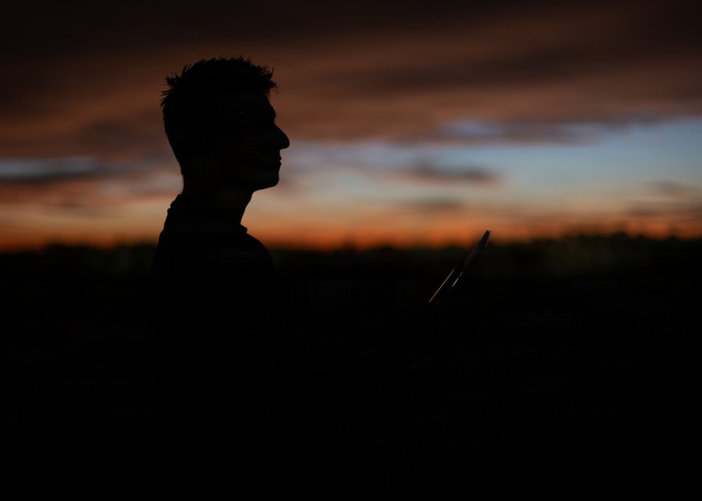 A silhouette of a person is captured against a dramatic sunset sky with orange, red, and blue hues. The figure appears to be holding a racket-like object, possibly a badminton or tennis racket, visible as a partial outline to the right of the subject. The image is intentionally dark with the subject rendered entirely in silhouette, creating a moody, artistic composition.