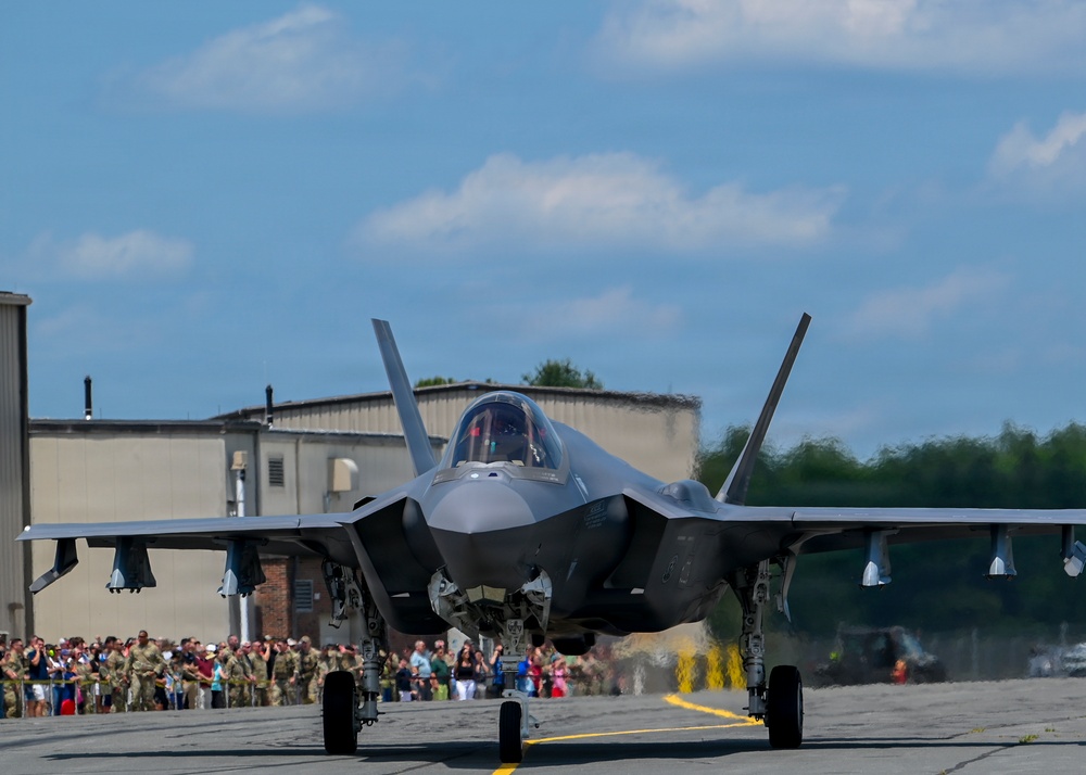 A Lockheed Martin F-35 Lightning II stealth fighter jet is taxiing on a runway apron during what appears to be a military air show. The aircraft is viewed head-on with its landing gear deployed, and a pilot is visible in the cockpit. A large crowd of spectators, including uniformed military personnel and civilians, lines the tarmac in the background.