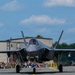 A Lockheed Martin F-35 Lightning II stealth fighter jet is taxiing on a runway apron during what appears to be a military air show. The aircraft is viewed head-on with its landing gear deployed, and a pilot is visible in the cockpit. A large crowd of spectators, including uniformed military personnel and civilians, lines the tarmac in the background.