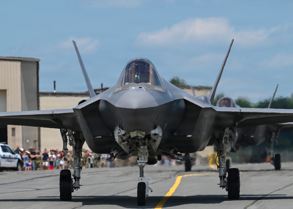 A front-facing view of an F-35 Lightning II stealth fighter jet taxiing on a runway apron, likely at a military air show. The aircraft is photographed head-on, showcasing its distinctive stealth design, canopy, and tricycle landing gear. A crowd of spectators and military personnel is visible in the background along with hangar structures.