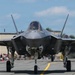 A front-facing view of an F-35 Lightning II stealth fighter jet taxiing on a runway apron, likely at a military air show. The aircraft is photographed head-on, showcasing its distinctive stealth design, canopy, and tricycle landing gear. A crowd of spectators and military personnel is visible in the background along with hangar structures.