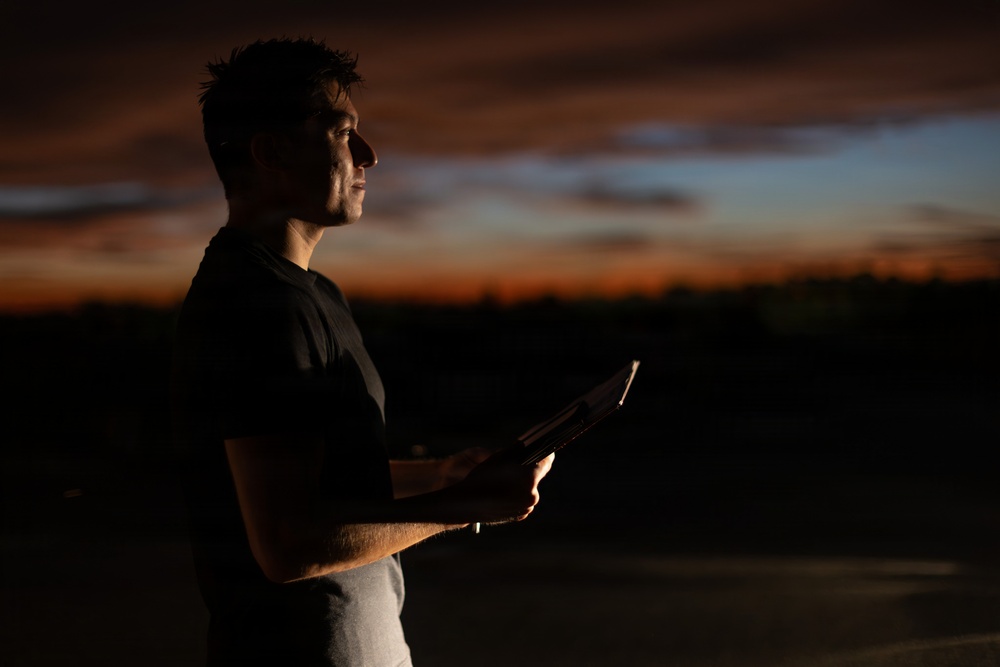 A young man stands outdoors during twilight or dusk, holding what appears to be a tablet or book that emits a faint light. He is photographed in profile against a dramatic sunset sky with deep orange, red, and blue tones. The image is artistically lit with strong contrast, leaving much of the subject in shadow.