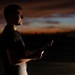 A young man stands outdoors during twilight or dusk, holding what appears to be a tablet or book that emits a faint light. He is photographed in profile against a dramatic sunset sky with deep orange, red, and blue tones. The image is artistically lit with strong contrast, leaving much of the subject in shadow.