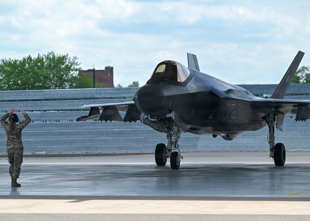 A military ground crew member in camouflage uniform is marshaling an F-35 Lightning II stealth fighter jet on a wet tarmac apron. The aircraft is taxiing with its landing gear deployed and cockpit canopy closed, with a pilot visible inside. The setting appears to be a military air base with hangar structures and trees visible in the background.