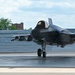 A military ground crew member in camouflage uniform is marshaling an F-35 Lightning II stealth fighter jet on a wet tarmac apron. The aircraft is taxiing with its landing gear deployed and cockpit canopy closed, with a pilot visible inside. The setting appears to be a military air base with hangar structures and trees visible in the background.