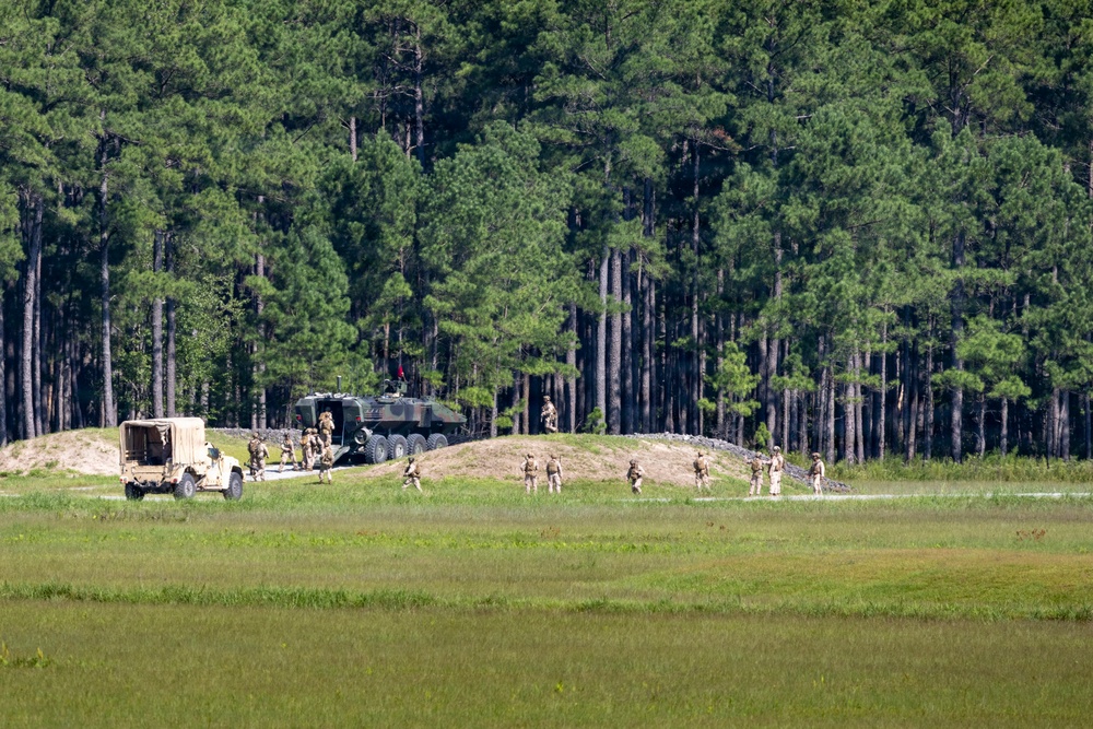 A military training exercise is taking place in an open field adjacent to a pine forest. Multiple soldiers in tan/desert camouflage uniforms are dispersed across a dirt road or path, with two military vehicles visible — a tan HMMWV (Humvee) and a larger green multi-axle tactical vehicle. The scene appears to depict a field training exercise with personnel dismounting or maneuvering around the vehicles.