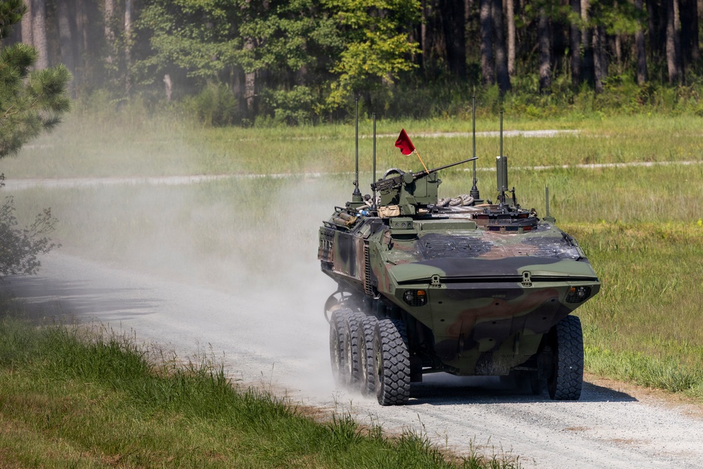 A wheeled armored military vehicle, consistent with a Light Armored Vehicle (LAV) or similar 8-wheeled APC, is traveling at speed along a gravel road kicking up a dust cloud. The vehicle is painted in green camouflage and is equipped with a mounted weapon system and multiple antennas. The setting appears to be a military training area surrounded by pine trees and open grassland.